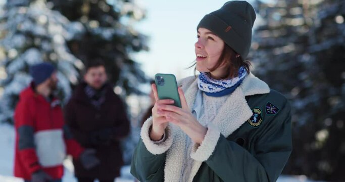 Happy excited young woman in winter outfit taking pictures on smartphone of beautiful scenery while staying with friends at winter resort.