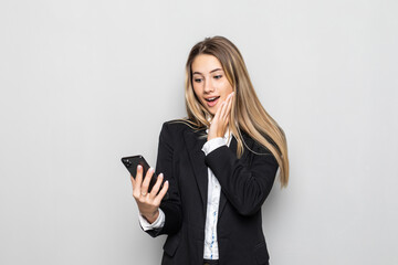 Fototapeta premium Portrait of surprised and impressed business woman with widened eyes and opened mouth, stunned on phone while standing against white background. Girl uses some app