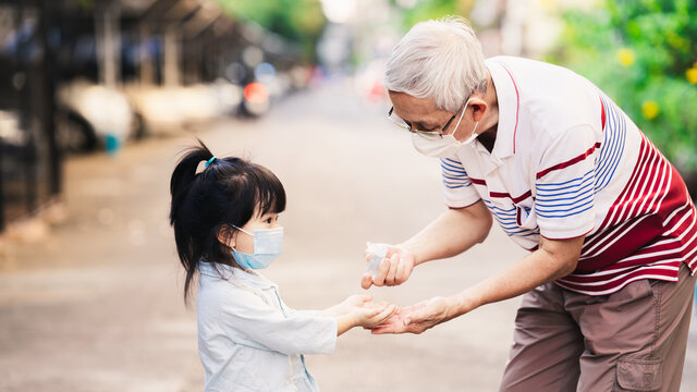 Family using alcohol gel washing hands. Grandfather squeezed alcohol gel on granddaughter to wash her hands. Adult and girl wear white and blue medical face masks. On winter season in Bangkok Thailand