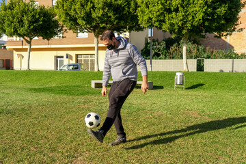 Young man playing soccer in a beautiful park with natural grass, wears a mask to protect himself from the coronavirus while doing sports. Copy space