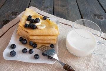 a mug with milk, next to blueberries, freshly baked pancakes poured with honey in a plate