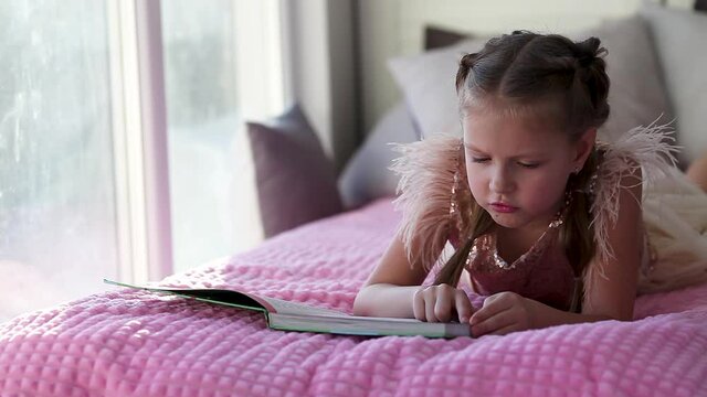 A little beautiful girl in a smart dress lies on the couch and reads a book.