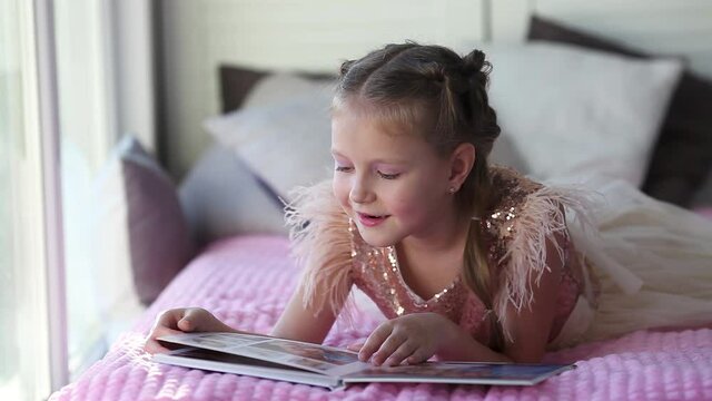 The little beautiful girl lies on the bed flips through a photo book from a family photo shoot . Beautiful and convenient storage of photos. Memory of an important period. Photo printing.