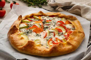 Homemade savory galette with vegetables, wheat pie with tomatoes, zucchini, blue cheese Gorgonzola. Rustic crust crostata on dark linen textile tablecloth, side view