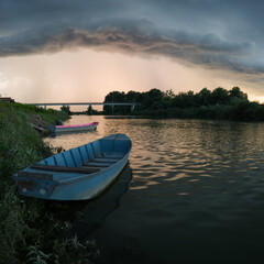 Storm arcus shaft and cumulonimbus cloud with heavy rain or summer shower, severe weather and sun glow behind rain. Landscape with Sava river with moored boats along grassy river bank and bridge.