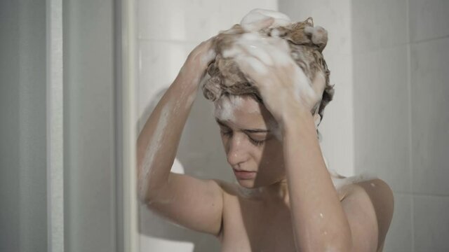 Portrait of Caucasian woman foaming shampoo standing in shower cabin. Adult slim lady taking douche in the morning at home. Body care and femininity concept.
