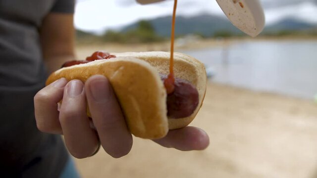putting ketchup on a hot dog or bratwurst during a picnic on the beach by a lake with mountain views.