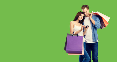 Love, holiday sales, shop, retail, consumer concept - happy couple with shopping bags, looking at mobile phone. Green color background.