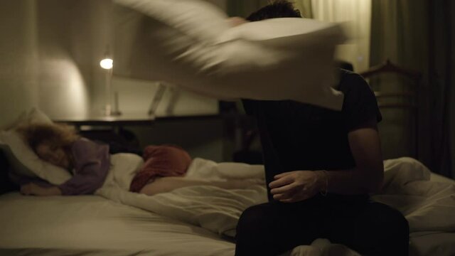 Stressed handsome man throwing pillow at snoring wife sleeping at background. Portrait of sad Caucasian husband sitting on bed at night with insomnia as wife sleep at home. Medical concept.
