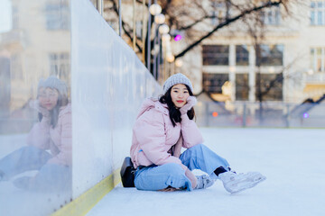 Ice skating concept. Fall on ice. Korean girl tying shoelaces on ice skates fell on ice in european city square, outdoor. Asian woman sitting on the ice rink smiling.