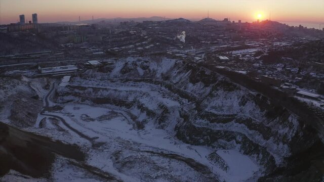 Aerial view of huge deep open pit covered with snow and Vladivostok cityscape is on the background. Evening. Russia