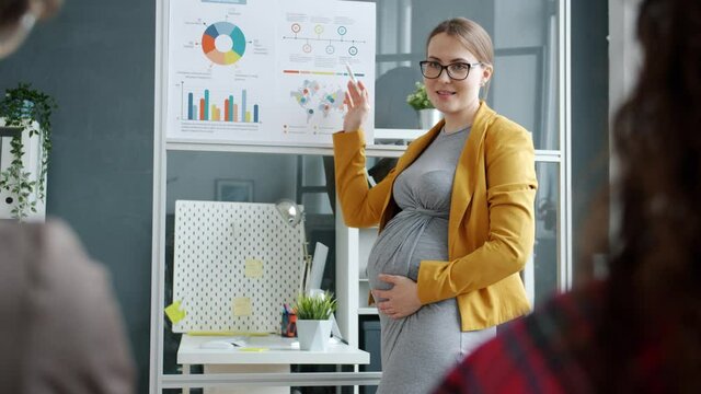 Ambitious employee pregnant girl is making presentation for people in office speaking pointing at glass board with charts discussing successful project