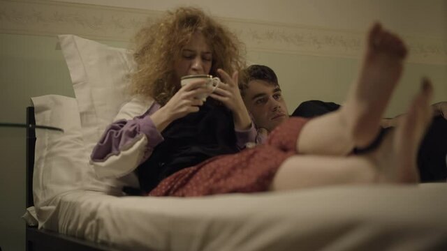 Relaxed husband and wife lying in bed watching TV as woman burning tongue with hot tea. Carefree Caucasian family weekend in bedroom at home. Marriage leisure concept.
