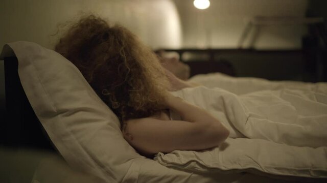 Side view of embarrassed woman waking up in bed naked with stranger man sleeping at background. Stressed Caucasian redhead lady awakening not at home. Hangover and lifestyle concept.