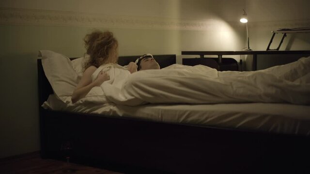 Wide shot of shocked woman waking up and checking naked man lying in bed. Wine glass standing on the floor. Embarrassed Caucasian lady lying in bedroom with stranger. Social concept.