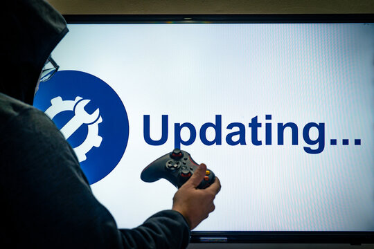 Hooded man holding a gaming controller in front of a screen showing an updating message for a game or software day 1 patch