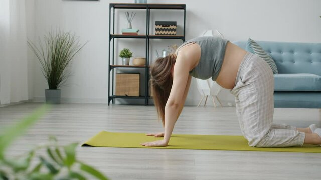 Slow motion of pregnant young woman in maternity wear doing yoga at home caring for body and baby exercising on yoga mat. People and wellness concept.