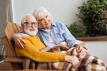 happy senior couple with plaid blanket looking at camera while hugging in armchair on blurred background on terrace