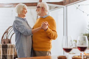 smiling elderly couple looking at each other while dancing with blurred wine glasses on foreground at home