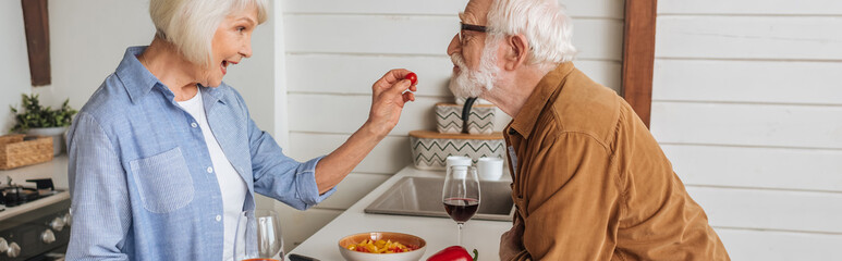side view of happy elderly wife feeding husband with cherry tomato in kitchen, banner