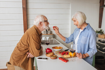 side view of happy senior wife feeding husband with cherry tomato near table with food, wine glasses and smartphone in kitchen