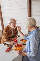 back view of senior wife feeding happy husband with cherry tomato near table with food in kitchen
