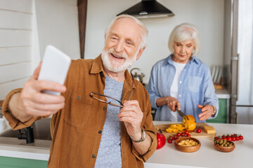 happy senior husband with smartphone taking selfie with wife cooking dinner in kitchen on blurred foreground