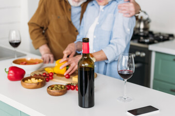 cropped view of elderly couple near table with bottle of wine and vegetarian food on blurred background