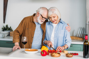 smiling senior husband hugging wife cooking dinner on table with wine, cheese and vegetables on blurred background
