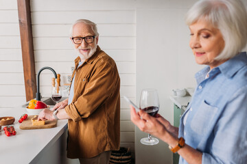 smiling senior man looking at woman while cutting cheese on chopping board in kitchen on blurred foreground