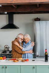 Fototapeta na wymiar happy elderly couple hugging near table with food and wine glasses in kitchen
