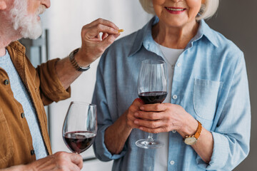 cropped view of elderly husband with glass of wine feeding wife with piece of cheese in kitchen