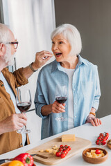 elderly husband with wine glass feeding happy wife with piece of cheese near table with food in kitchen on blurred foreground