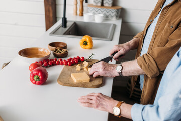 cropped view of elderly wife near husband cutting cheese on chopping board in kitchen on blurred background