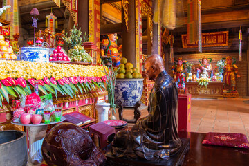 Polished bronze statue of a monk kneeling in front of richly decorated altar with harvested food inside a Buddhist temple atop Mount Fansipan near Sapa in northern Vietnam