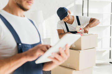 indian mover packing box near coworker holding digital tablet on blurred foreground