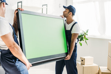 multicultural movers in caps and uniform carrying plasma tv with green screen in apartment