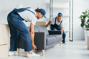 happy multiethnic movers in uniform smiling while carrying couch in apartment