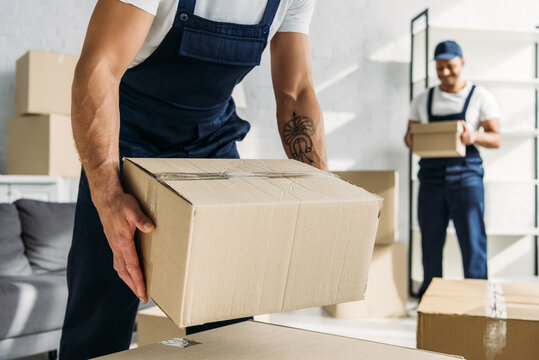 cropped view of tattooed worker in uniform holding carton box near indian coworker