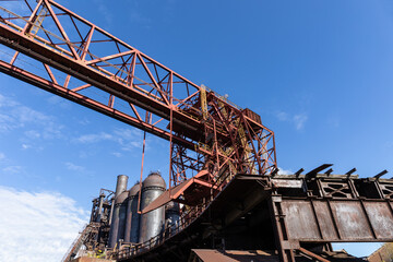 Old steel mill with overhead trusswork, rusting blast furnaces against a blue sky, horizontal aspect