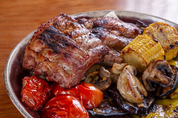 Colorful grilled vegetables served with meat on red plate and resting on wooden table surface with copy space. BBQ party with friends.