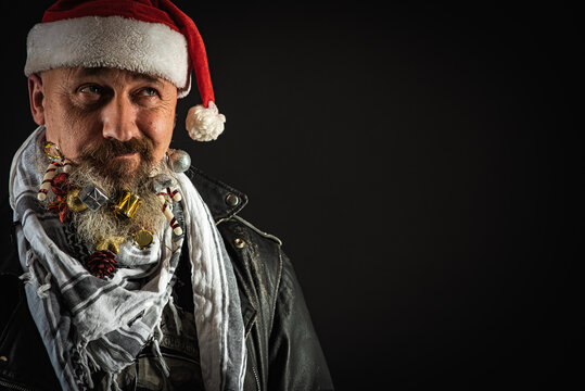 a young, handsome, serious man with a long beard on which hang Christmas toys, a man in a black jacket and a Christmas hat on his head on a dark background, emotional. There is a place for an inscript