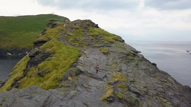 Hiker climbing and ascending rocky coastal headland cliff, Cornwall UK, aerial