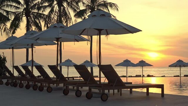 A colorful sunset highlights a line of empty sun loungers and beach umbrellas along the edge of a resort swimming pool.