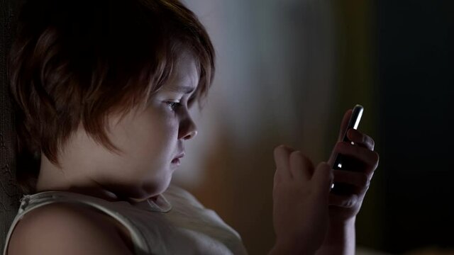 Portrait in profile of a small girl with short hair and plump cheeks, in a t-shirt lying at home. She holds in her hands and uses a smartphone for games.