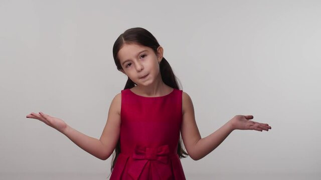 Surprised puzzled puzzled little girl in red dress posing in the studio isolated on white color background. His hands point to both sides.