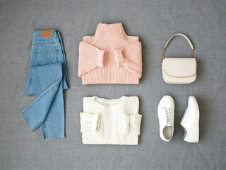 Blue jeans, pink and white knitted sweaters, small cross body bag and sneakers on grey background. Overhead view of woman's casual outfit. Trendy stylish look. Women clothes. Flat lay, top view.