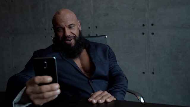 a muscular, bald, bearded man in a black shirt sits at a Desk and uses a mobile phone, talking on a video link and smiling.