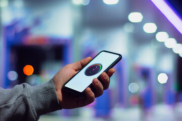 A man holds a smartphone with a digital fuel meter on the screen against the background of a night gas station for a car.