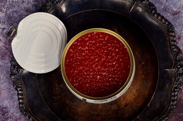 Open tin can with red salmon caviar on antique silver plate. Rustic background, top view, copy space